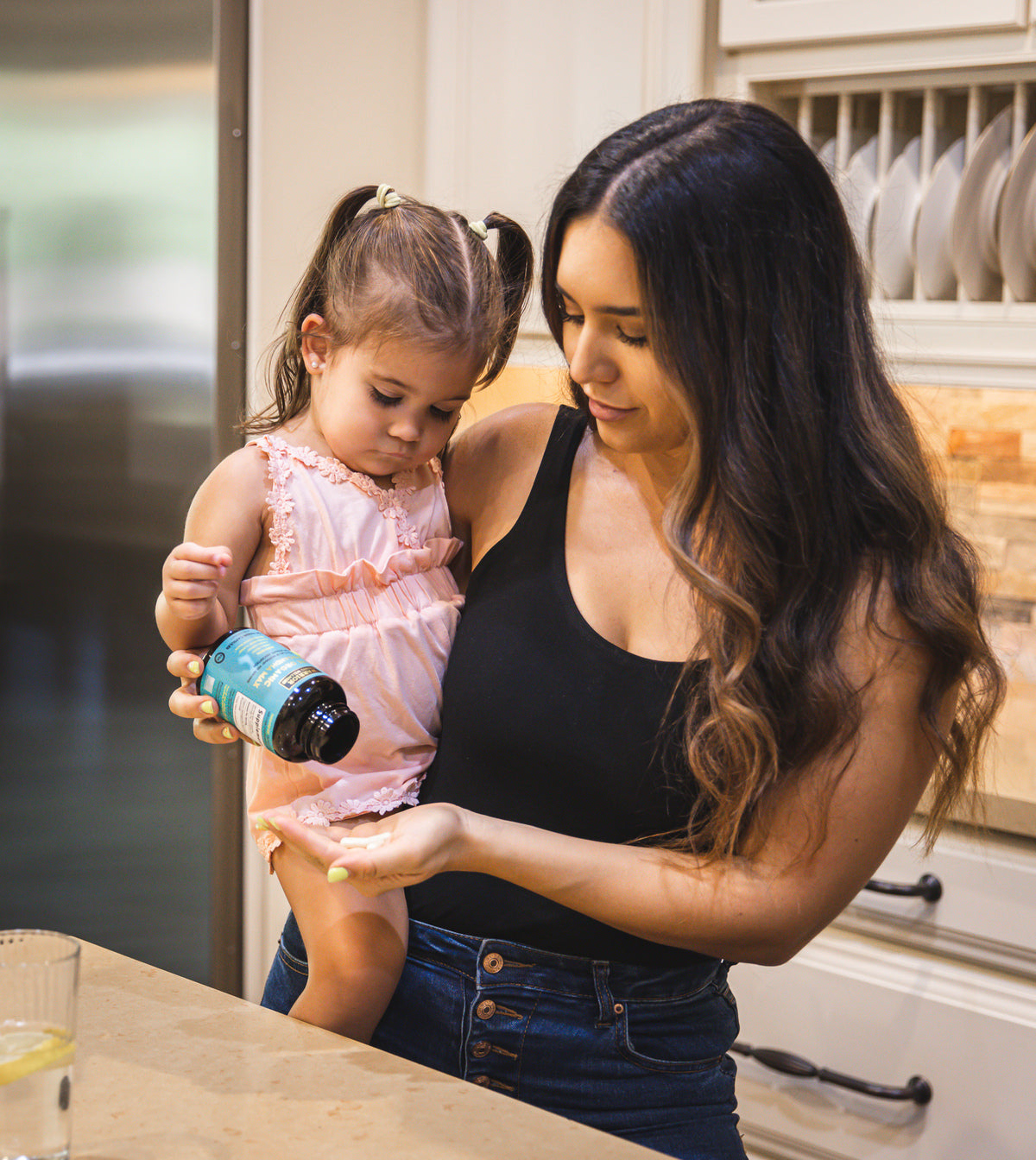Mother taking Ashwagandha supplement while holding her daughter on her hip in a kitchen.
