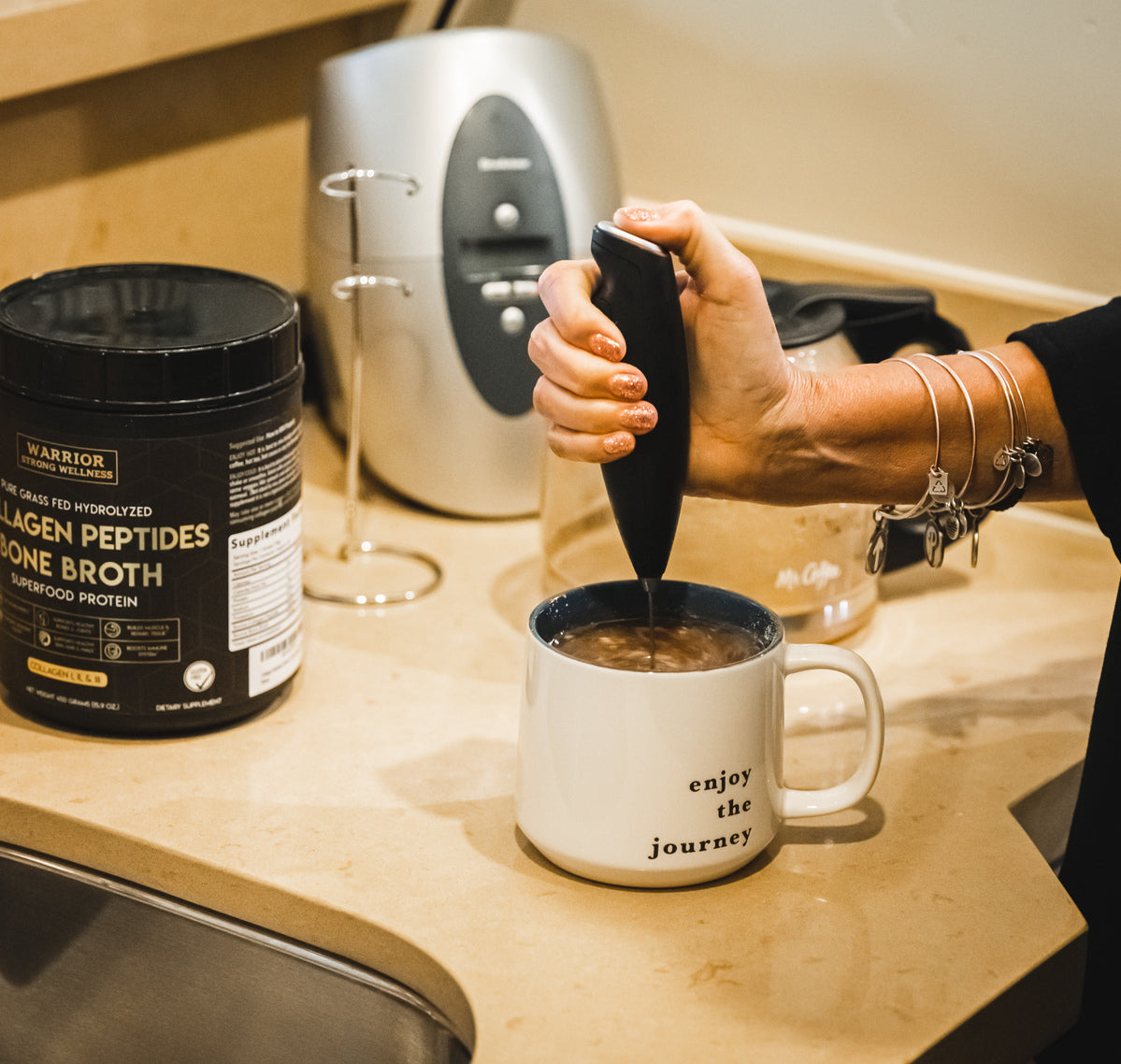 Handheld milk frother blending collagen powder into coffee beside Warrior Strong Wellness Collagen Peptides & Bone Broth, demonstrating easy mixing for drinks and recipes.