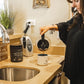 Woman preparing coffee with Warrior Strong Wellness Collagen Peptides & Bone Broth, showcasing an easy way to add collagen protein to a daily morning routine.