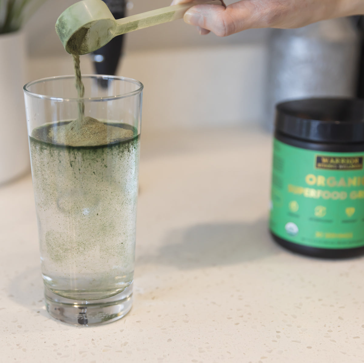 Organic Superfood Greens powder being added to water with a scoop, demonstrating easy daily mixing before enjoying a nutrient-rich greens drink.